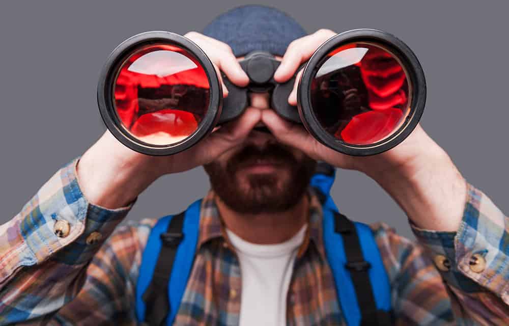 A man in a plaid shirt and beanie hat looks through over-sized binoculars, directly at us.