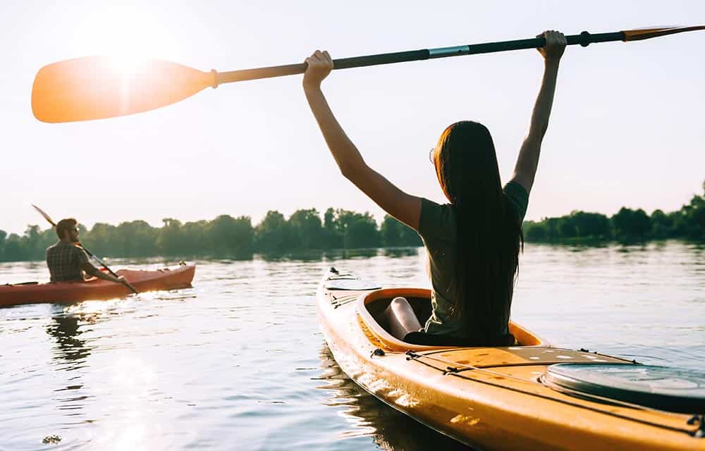 Two kayakers crossing a still expanse of water. The female kayaker is holding her paddle aloft in a triumpant gesture.